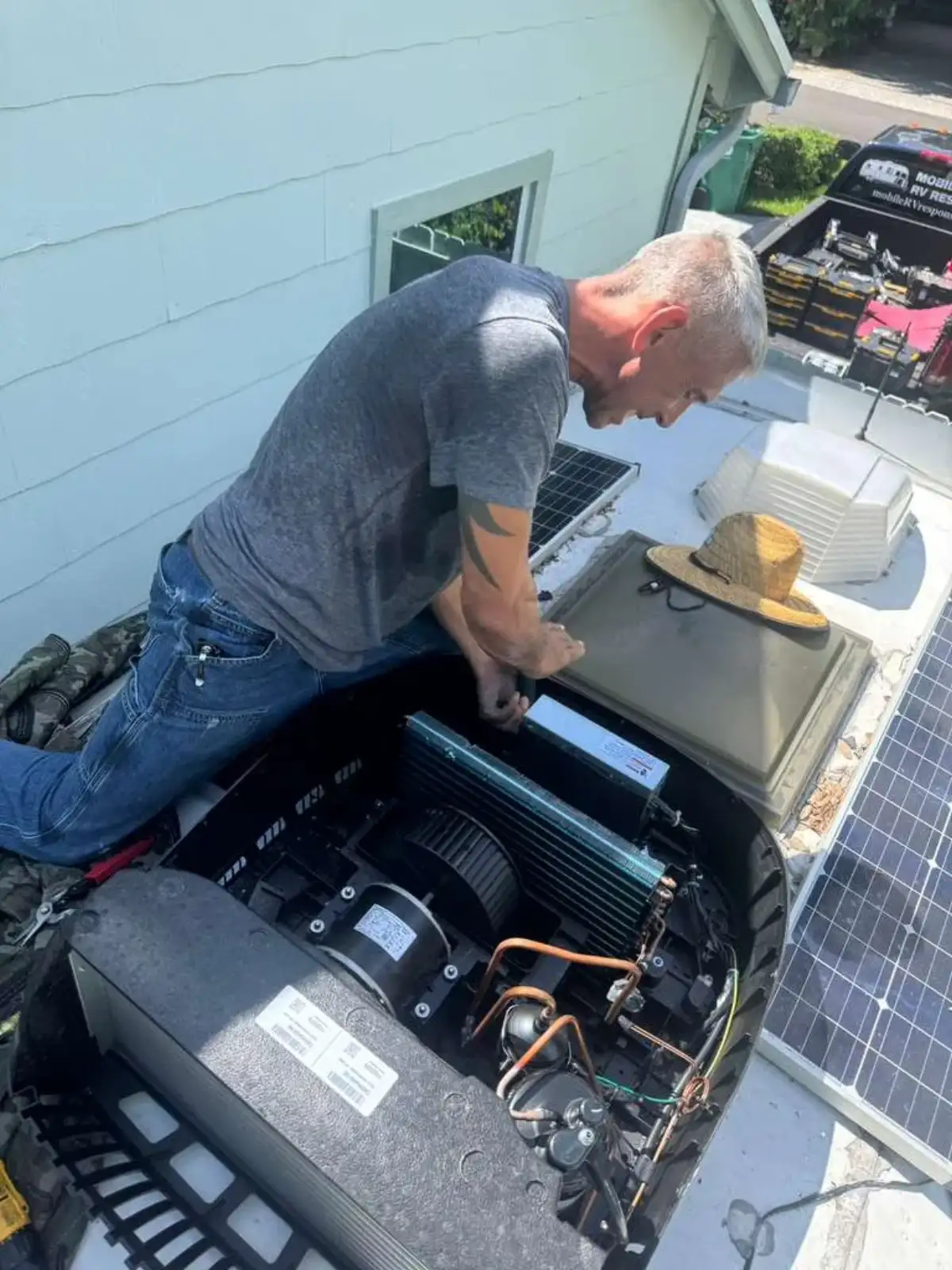 Technician kneeling on an RV roof beside solar panels with both hands inside an opened rooftop air conditioner