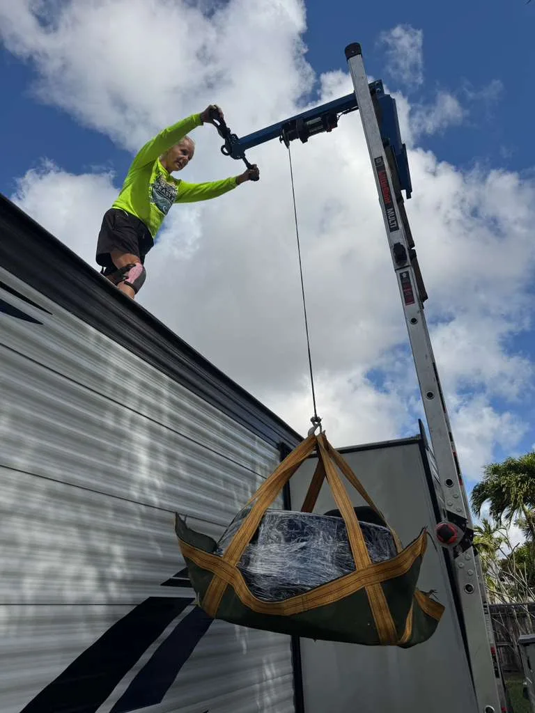 Technician on an RV roof hoisting a shrink-wrapped replacement air conditioner in a sling from a ladder hoist under Florida palms