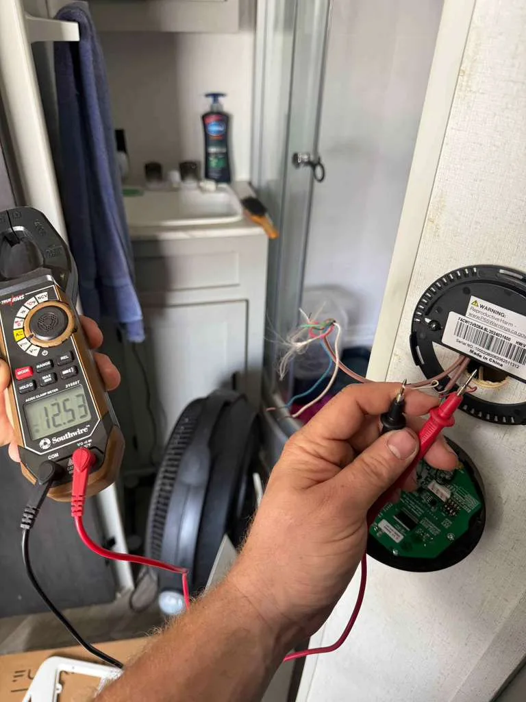 Hand holding a multimeter reading 12.55 volts across a control board pulled from an RV wall